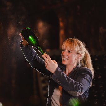 Eine blonde Frau in einem Weinkeller hebt eine Flasche, während sie von weiteren Flaschen umgeben ist und lächelt.