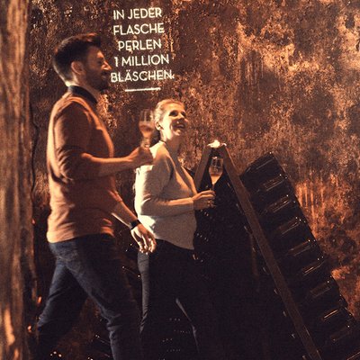 Ein Paar genießt einen entspannten Moment mit Sekt in der Hand, umgeben von einer rustikalen Atmosphäre und Flaschenregalen.