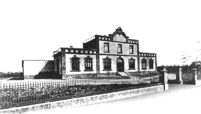 Ein historisches Gebäude mit einer markanten Fassade und großen Fenstern, umgeben von einem niedrigen Zaun.