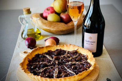 Der Zwiebelkuchen, reich belegt mit Oliven und Sardellen, wird von einer Flasche Geldermann Rosé und frischen Äpfeln umrahmt.