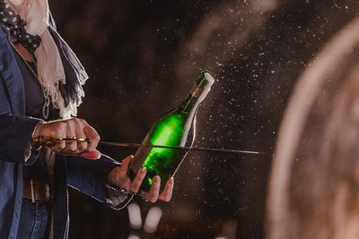 Eine gruene Flasche, die geöffnet ist und aus der Schaum fließt, im Hintergrund ein Holzfaß und Arm sowie Hand einer Frau 
