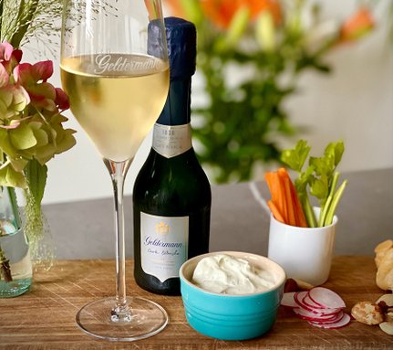 Ein elegantes Glas Sekt steht neben einer Flasche Geldermann, umgeben von frischem Gemüse und einer cremigen Dip-Schale.