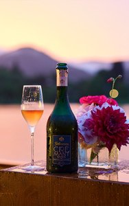 Geldermann Soirée Rosé: Eine Flasche Geldermann Crémant Baden und ein Glas Rosé Sekt vor der Kulisse des Tegernsees im Abendrot