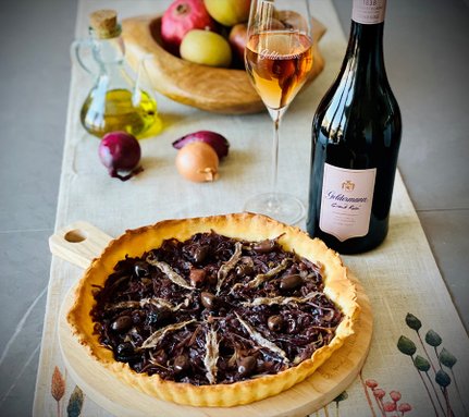 Der Zwiebelkuchen, reich belegt mit schwarzen Oliven und Sardellen, steht auf einem Tisch mit frischen Äpfeln und Zwiebeln.