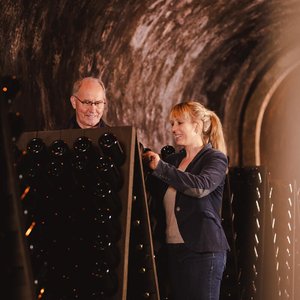 Ein älterer Mann und eine Frau arbeiten gemeinsam in einem Weinkeller, um Flaschen auf einem Gestell zu sortieren.