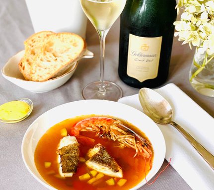 Ein elegantes Gericht mit Fisch und Garnelen in einer aromatischen Brühe, serviert mit frischem Brot und einem Glas Sekt.