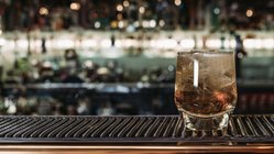 Das Glas mit Eis und einem erfrischenden Getränk reflektiert das Licht, während die Bar im Hintergrund eine lebhafte Atmosphäre schafft.