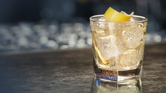 Das Getränk im Glas sprudelt leicht, während ein Zitronenstück dekorativ darauf liegt, umgeben von einer unscharfen Bar-Umgebung.