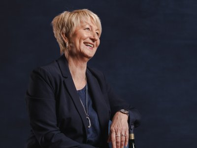 Die Frau trägt einen eleganten Anzug und zeigt eine freundliche Ausstrahlung, während sie auf einem Hocker sitzt.