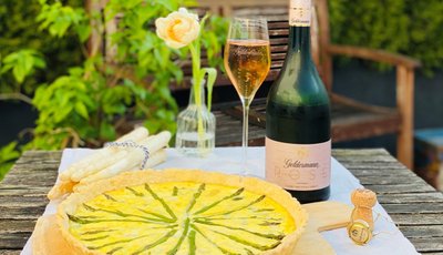 Ein rustikaler Tisch mit einem Spargelkuchen, einem Glas Rosé und einer Flasche Sekt, umgeben von grünen Pflanzen.