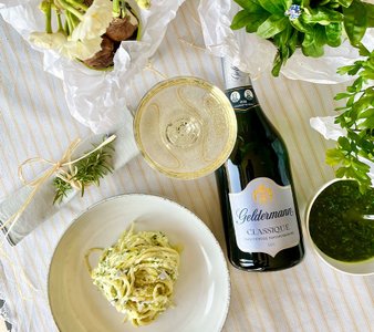 Ein eleganter Tisch mit einem Teller Pasta, einem Glas Sekt und frischen Kräutern, umgeben von blühenden Pflanzen.