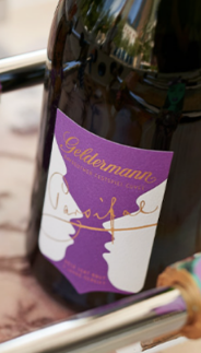 Auf einem Tisch präsentiert sich eine Flasche Geldermann Sekt, flankiert von bunten, stilisierten Figuren, die das Ambiente auflockern.