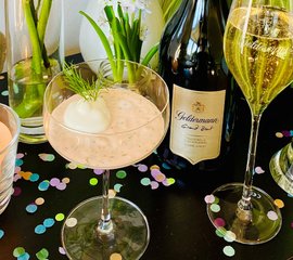 Das Bild zeigt ein Glas mit einer schaumigen Speise, umgeben von Sekt und bunten Dekorationen auf einem dunklen Tisch.