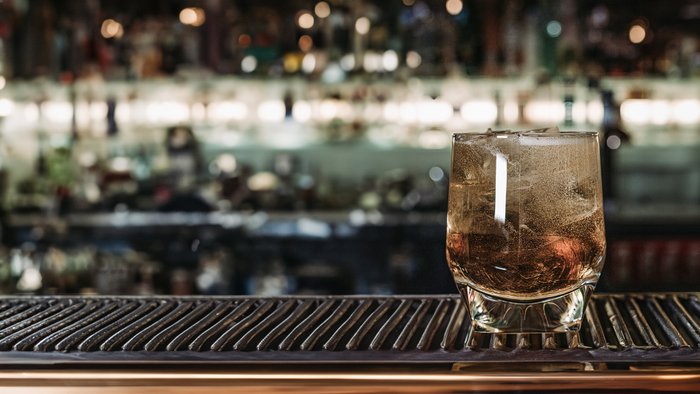 Das Glas mit Eis und einem erfrischenden Getränk reflektiert das Licht, während die Bar im Hintergrund eine lebhafte Atmosphäre schafft.