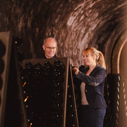 Ein älterer Mann und eine Frau in einem Weinkeller, die zusammen an Flaschen arbeiten, strahlen Freude und Teamgeist aus.