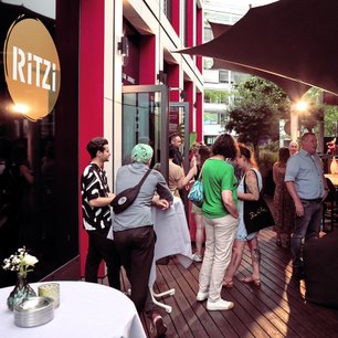 Eine Gruppe von Menschen steht vor einem Restaurant mit dem Namen 'RITZi', während sie sich unterhalten und lachen.