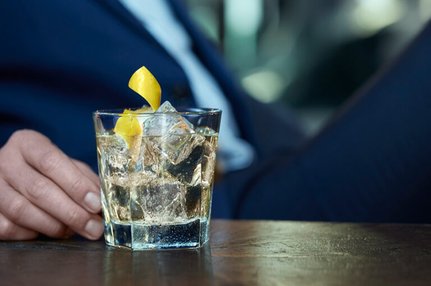 Eine Hand in einem Anzug hält ein Glas mit Eis und einem gelben Zitronenstreifen, während im Hintergrund unscharfe Farben zu sehen sind.
