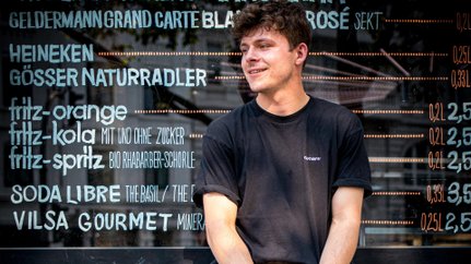 Der Mann trägt ein schwarzes T-Shirt und kurze Hosen, während er lächelnd auf einer Erhöhung sitzt, die mit Graffiti verziert ist.
