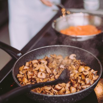 In einer Pfanne braten sich goldbraune Champignons, während Dampf aufsteigt und ein Koch im Hintergrund aktiv ist.