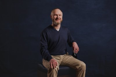 Mit einem freundlichen Lächeln sitzt der Mann auf einem Hocker und hält eine Flasche, umgeben von einem tiefblauen Hintergrund.