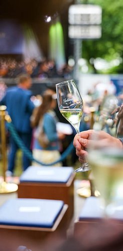 Ein Sektglas mit der Bayreuther Festspiel Cuvée 2025 vor dem Hintergrund der Open-Air-Aufführung während der Bayreuther Festspiel