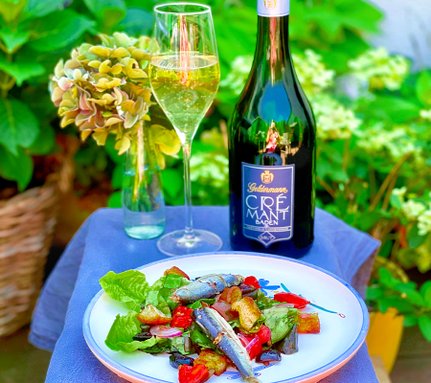 Ein frischer Salat mit Sardinen, serviert auf einem Teller, umgeben von üppigem Grün und einer Flasche Crémant.