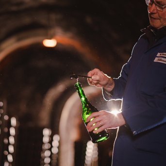 Der Keller ist mit vielen Flaschen gefüllt, während ein Mann konzentriert eine Flasche in der Hand hält und sie öffnet.