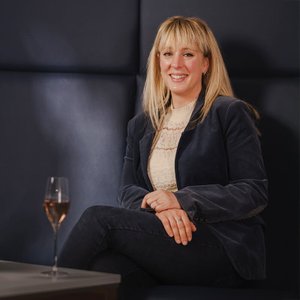 In einem geschmackvoll eingerichteten Raum sitzt die Frau mit blonden Haaren, die in einem Blazer und einer Bluse gekleidet ist.