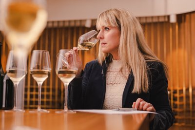 Eine Frau mit langen blonden Haaren hält ein Weinglas und betrachtet es nachdenklich, während sie an einem Tisch sitzt.