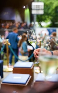 Ein Sektglas mit der Bayreuther Festspiel Cuvée 2025 vor dem Hintergrund der Open-Air-Aufführung während der Bayreuther Festspiel