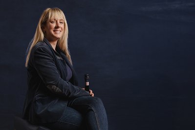 Mit einem selbstbewussten Lächeln sitzt die blonde Frau auf einem Stuhl, ihre Körperhaltung strahlt Gelassenheit aus.