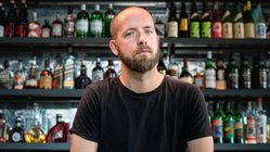 Der Bartträger in einem schwarzen T-Shirt wirkt nachdenklich, während er auf einer Bar sitzt, die mit Flaschen dekoriert ist.