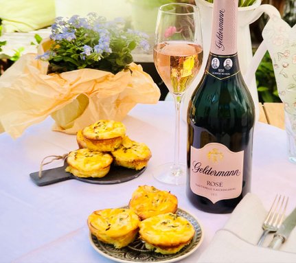 Ein romantisches Picknick-Setting mit Roséwein, einer Flasche Geldermann und köstlichen Quiches, umgeben von blühenden Pflanzen.