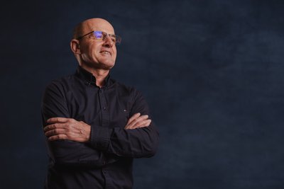 Der selbstbewusste Mann in einem schwarzen Hemd hat die Arme vor der Brust verschränkt und blickt nachdenklich zur Seite.