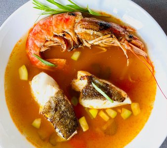 Die Suppe enthält eine große, rote Garnele und zwei zarte Fischstücke, umgeben von einer würzigen Brühe und Gemüsewürfeln.