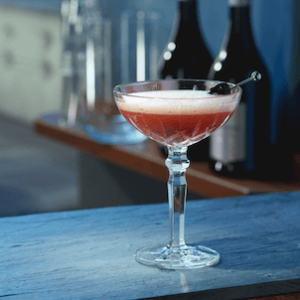 Cocktailschale mit hellrotem Sektcocktail, Schaumkrone und Amarena-Kirsche auf einem Tresen.