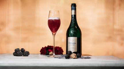 Das Glas mit dem prickelnden Getränk ist mit Wassertropfen bedeckt, umgeben von Brombeeren und roten Blumen.
