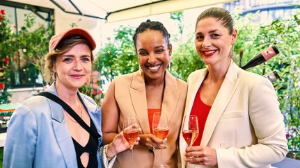 Mit Gläsern in der Hand posieren drei Frauen in modischen Outfits vor einer blühenden Kulisse und genießen den Moment.